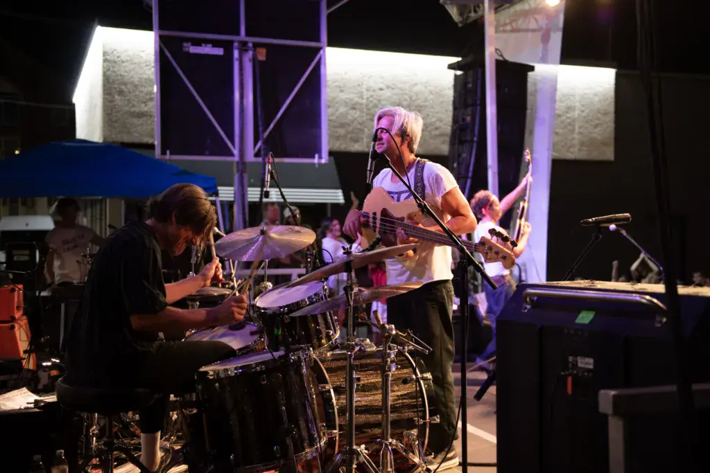A band performs on a lit stage at night, with a drummer and guitarist visible among the band members.