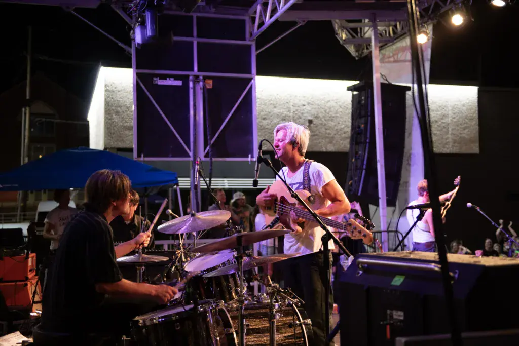 Musicians perform on an illuminated stage at night, with a drummer in foreground and guitarist singing at the microphone.