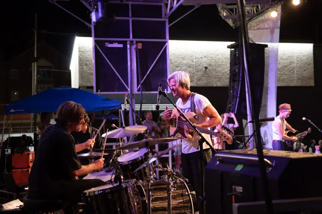 A band performs on an illuminated outdoor stage at night, with musicians playing drums, bass, and guitar.