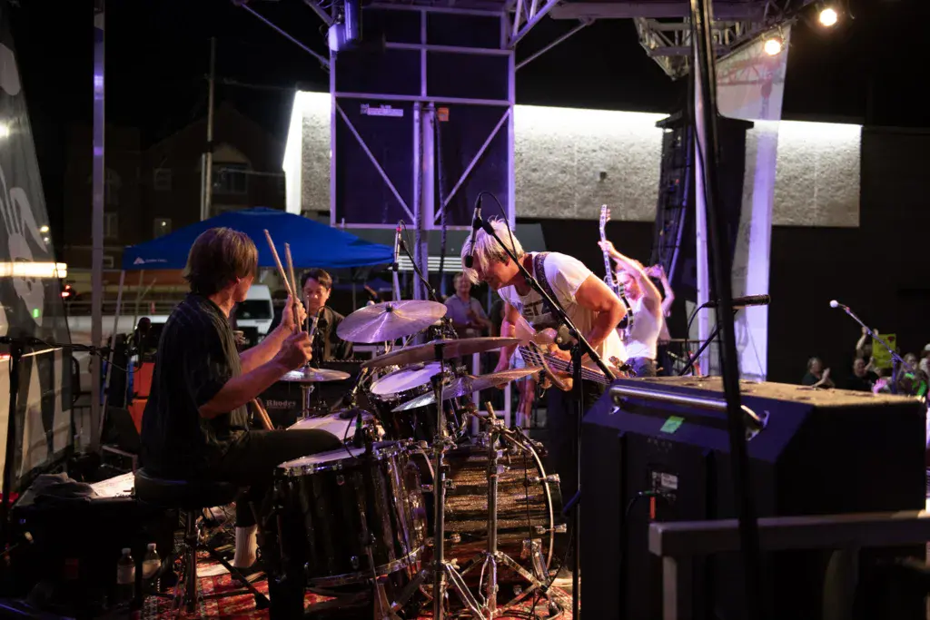 Band performing on an illuminated outdoor stage at night with drummer in foreground and other musicians visible.