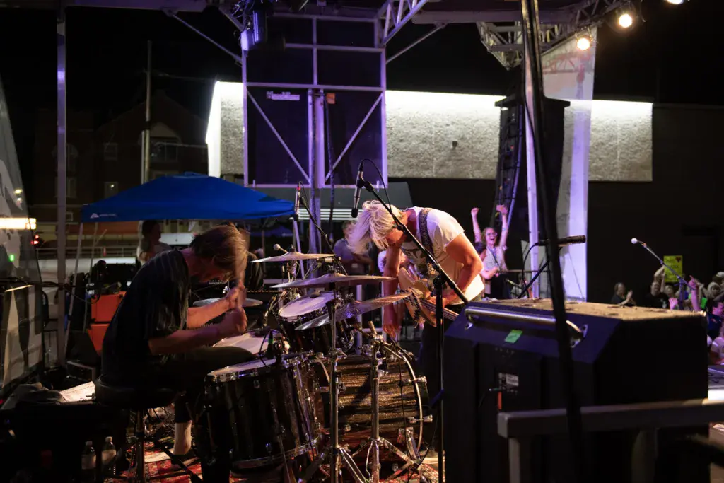 Drummer performs on stage under purple lighting at an outdoor nighttime music festival.