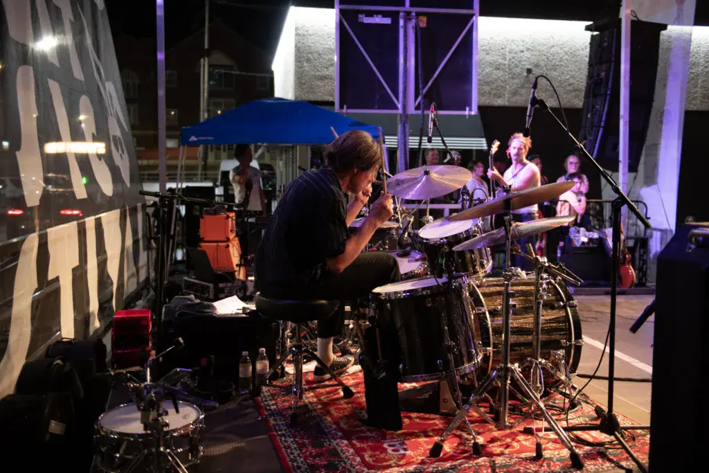 A drummer performs on stage at an outdoor festival venue under purple lighting at night.