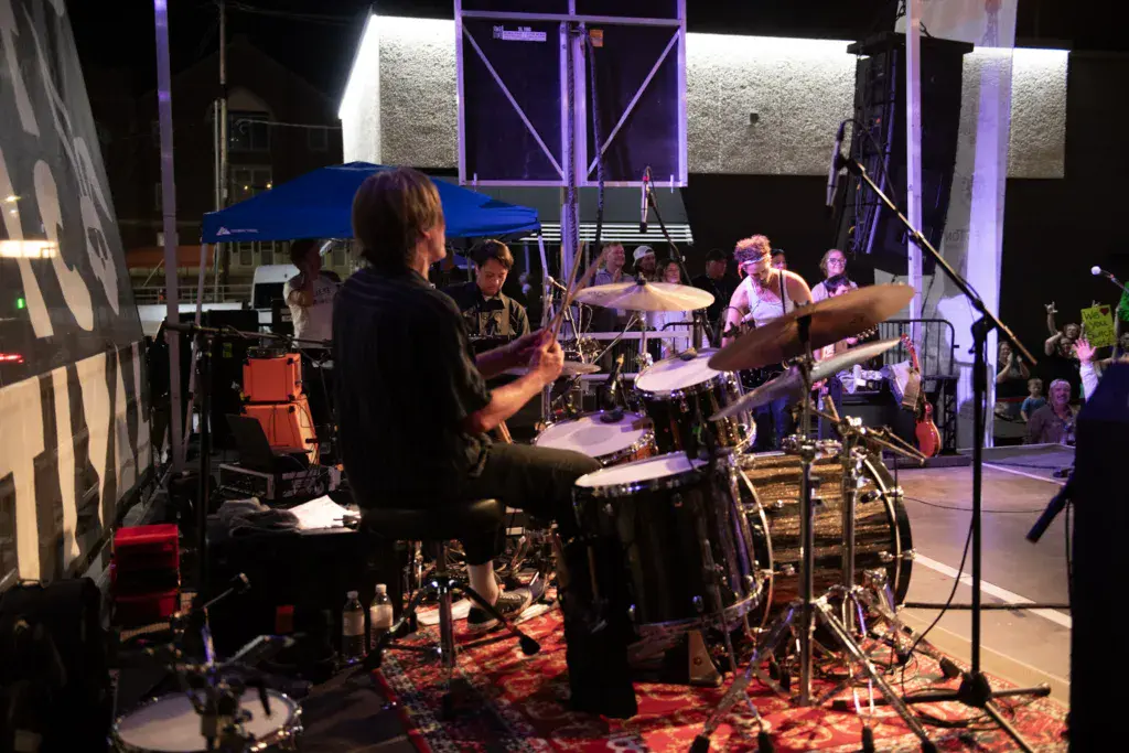 A drummer performs on stage at an outdoor music festival with a full band and audience visible in the background.