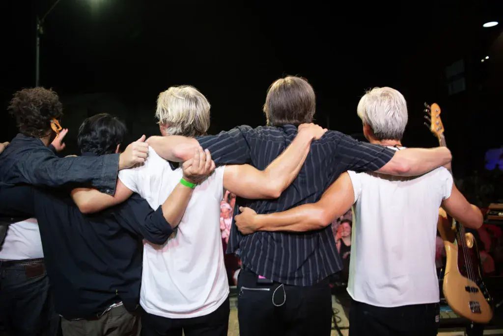 A group of band members and associates stand together with their arms around each other on stage at night.