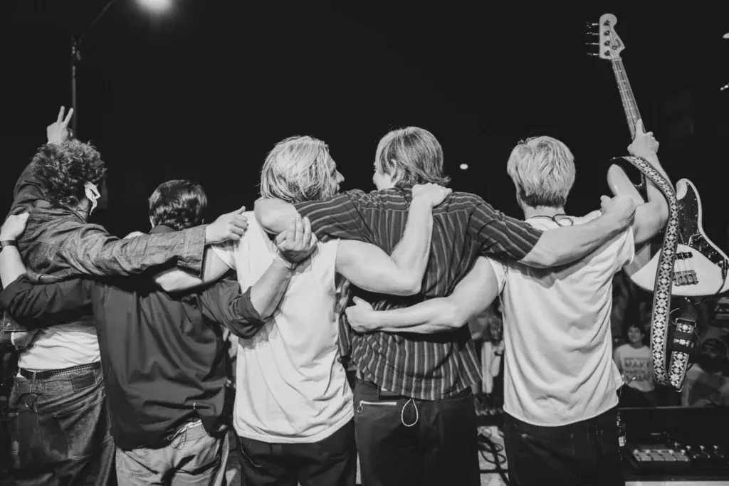 Band members on stage with arms around each other in a moment of unity during a performance.